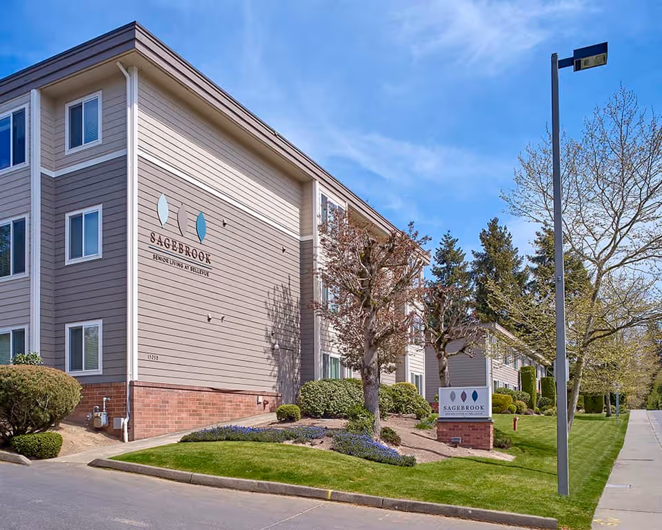 Front exterior of the Sagebrook Senior Living at Bellevue building with landscaped lawn and a sign under a blue sky.