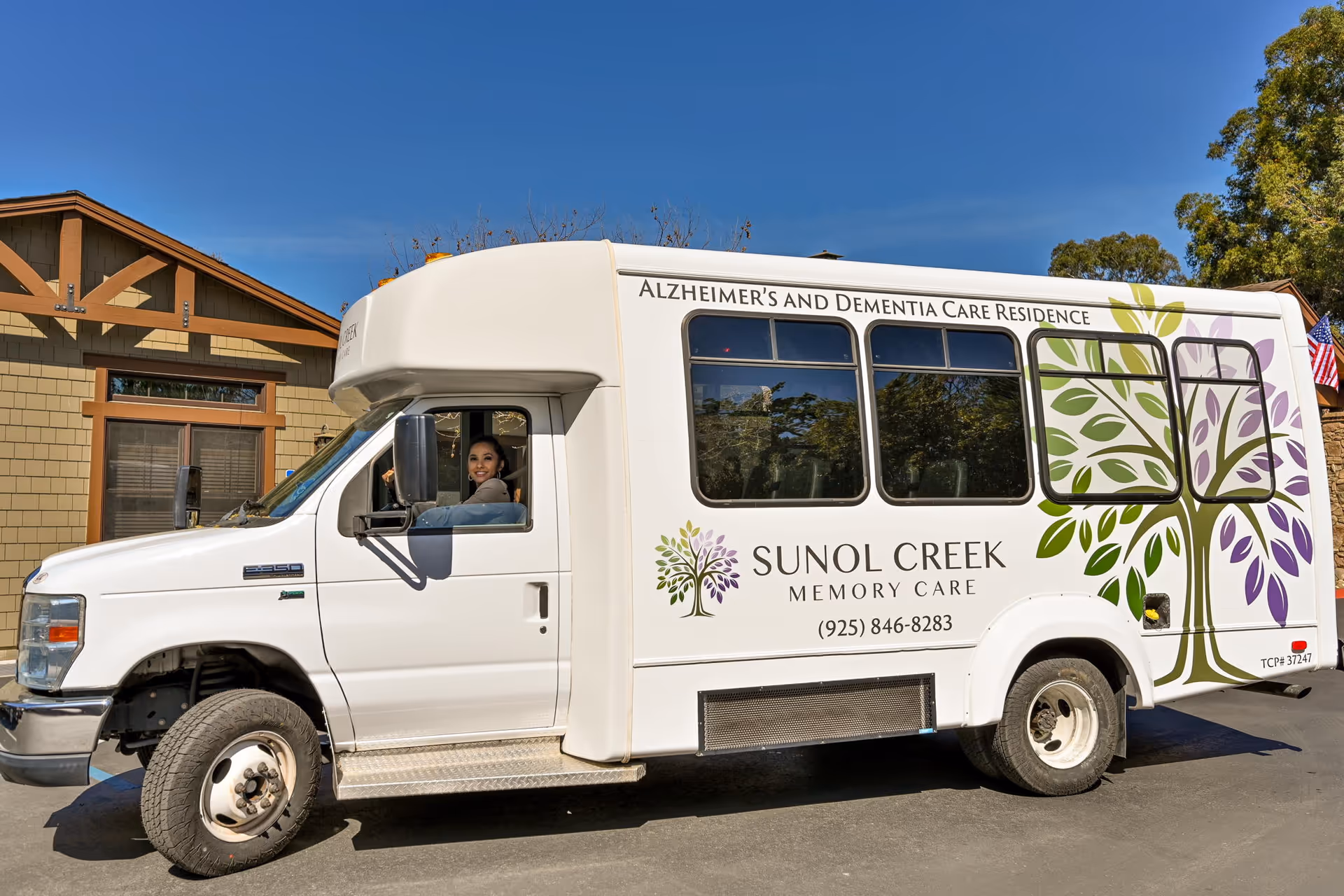 A white Sunol Creek Memory Care shuttle bus with a tree graphic parked outside a building, a woman visible in the driver's seat.