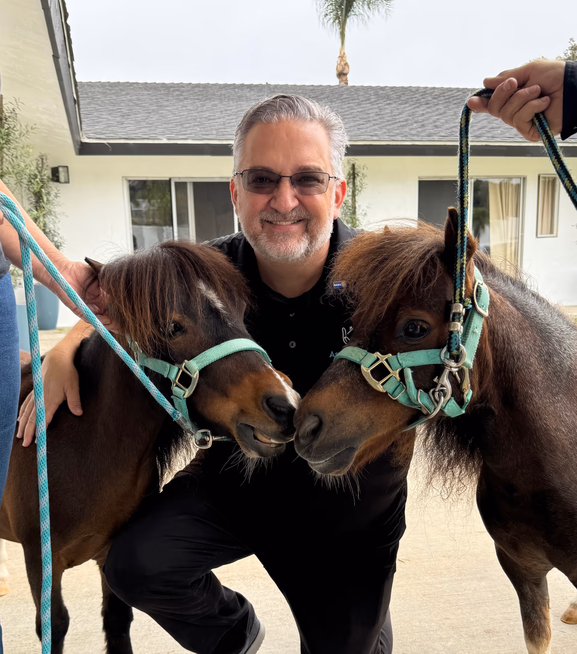 Smiling man with two ponies outdoors at a senior living campus.