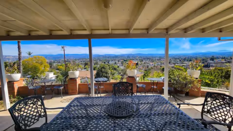 Covered outdoor patio with metal table and chairs overlooking a cityscape and distant mountains, with potted plants on a brick railing.