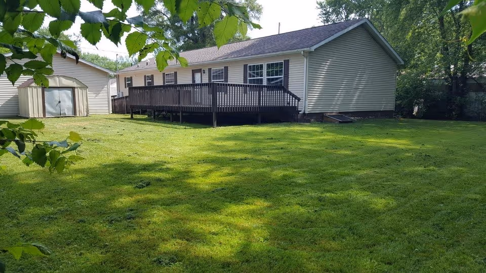 A single-story building with a wooden deck on a grassy, tree-shaded lawn.