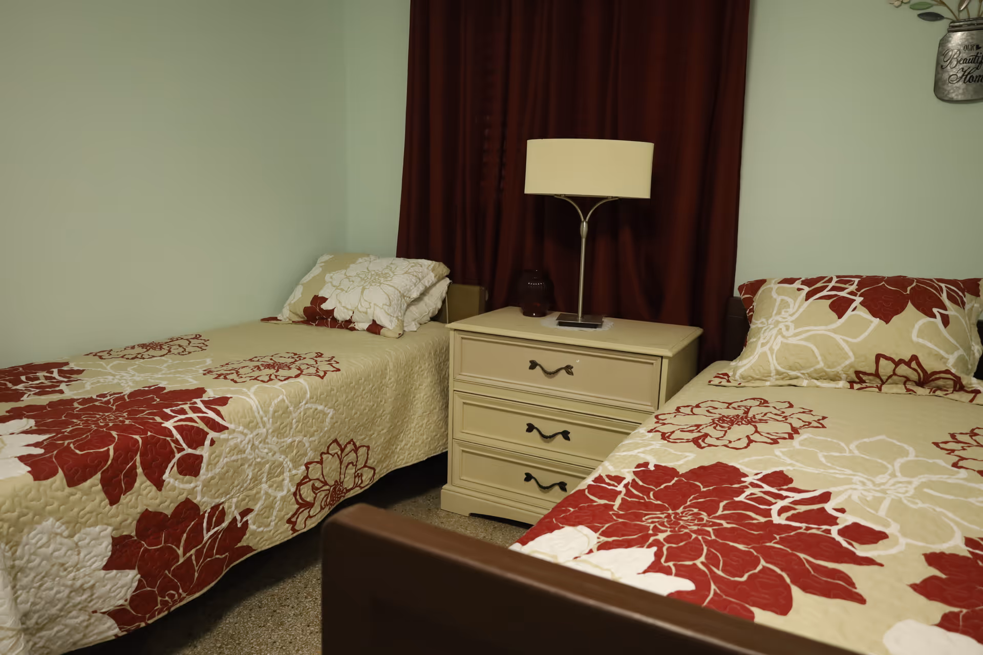 Two twin beds with floral quilts flank a nightstand and lamp in a pastel bedroom.