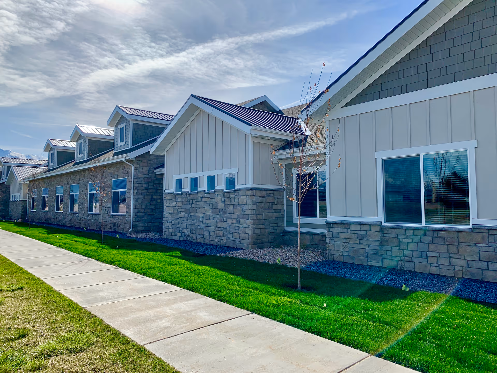 Exterior view of a single-story assisted living building with stone and siding facade, multiple windows, young trees, and a sidewalk.