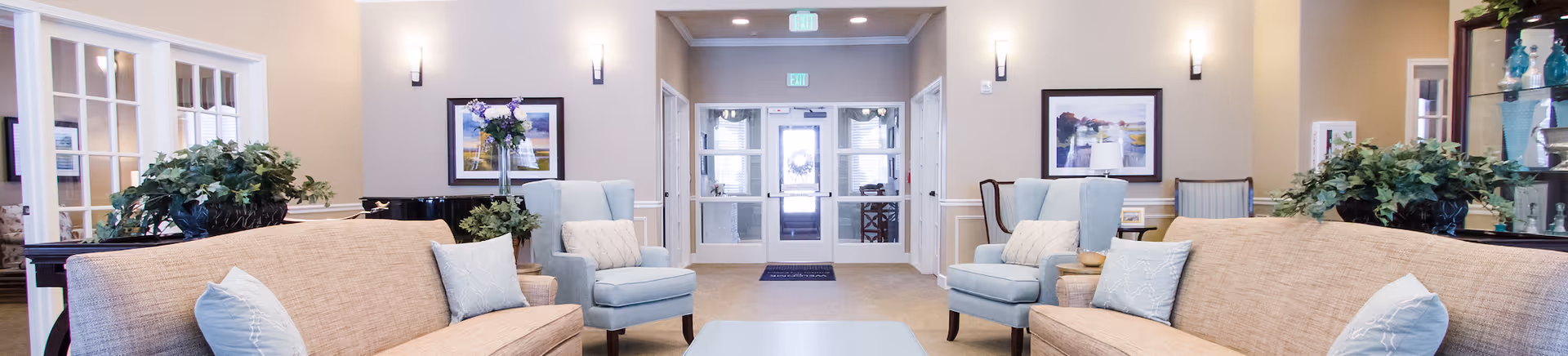 Bright, welcoming lobby seating area with sofas, armchairs, a coffee table and plants facing double entrance doors.
