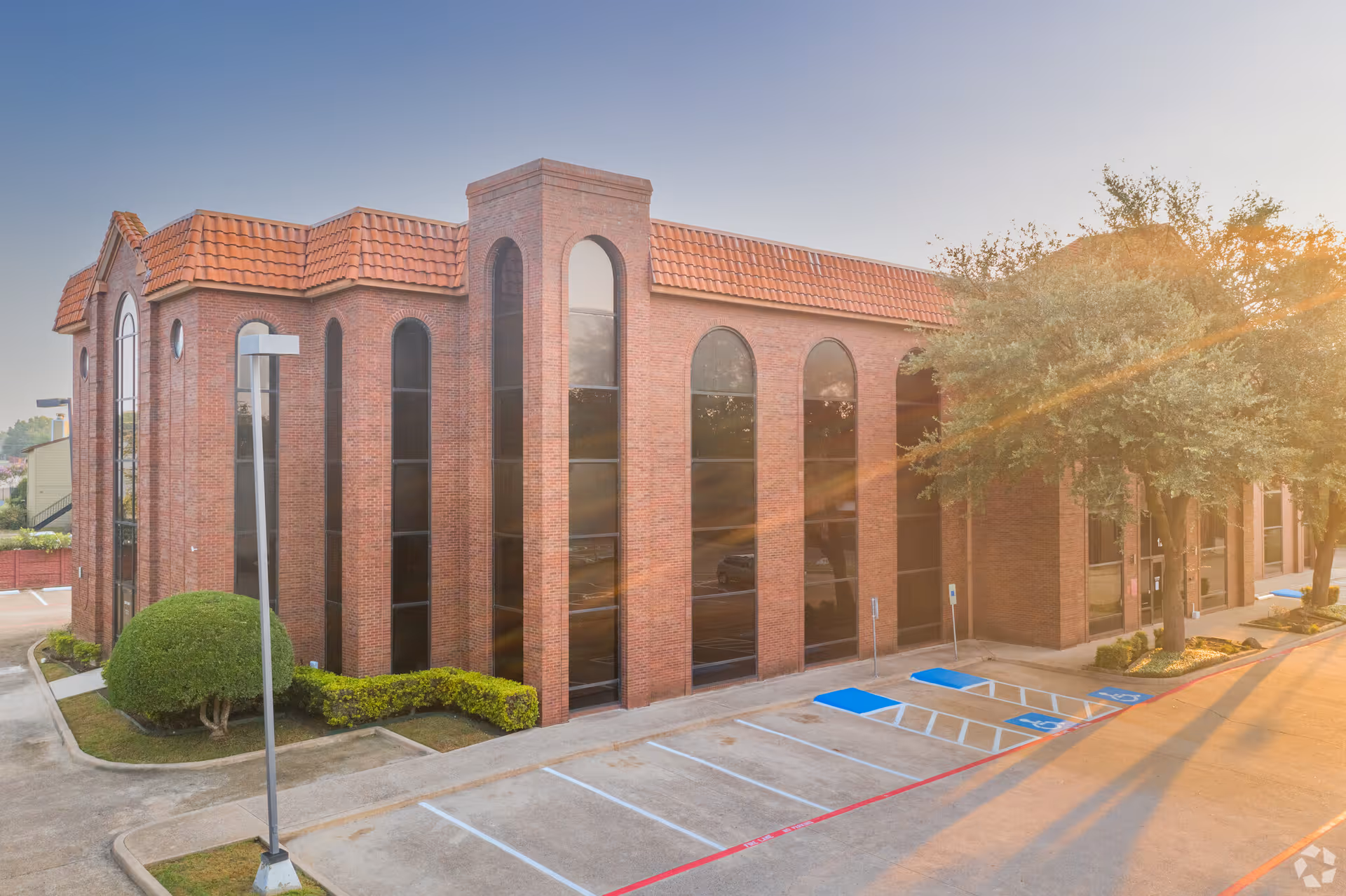 Brick building with arched windows, a parking lot, trees, and sunlight.