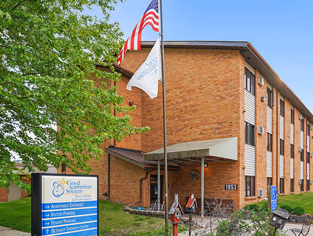 Brick senior living building with entrance canopy, flags, and a Good Samaritan Society sign.
