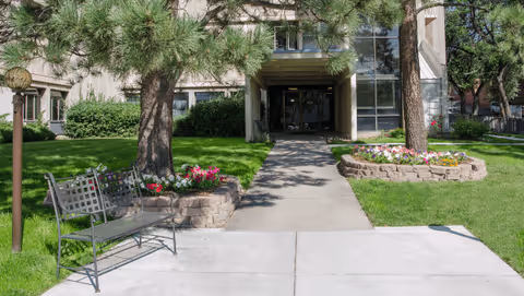 Front entrance of a building with a paved walkway leading to glass doors, flanked by trees, flower beds, and a bench.