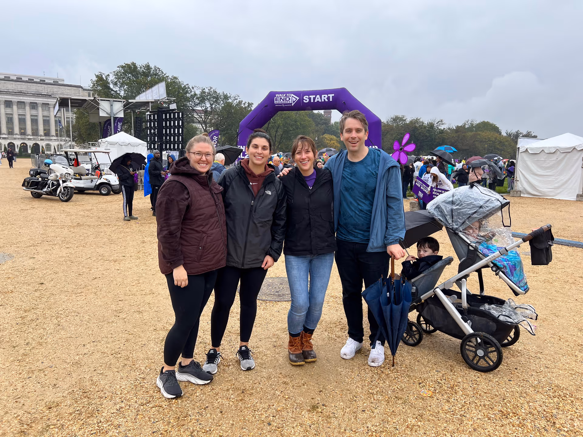 Group of five adults posing outdoors at a start line with a stroller