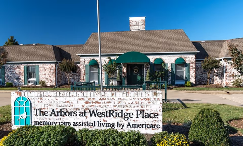Exterior view of The Arbors at WestRidge Place senior living facility with a brick building, green window shutters, and a green awning over the entrance. A sign in front reads 'The Arbors at WestRidge Place memory care assisted living by Americare' surrounded by landscaping and bushes under a clear blue sky.