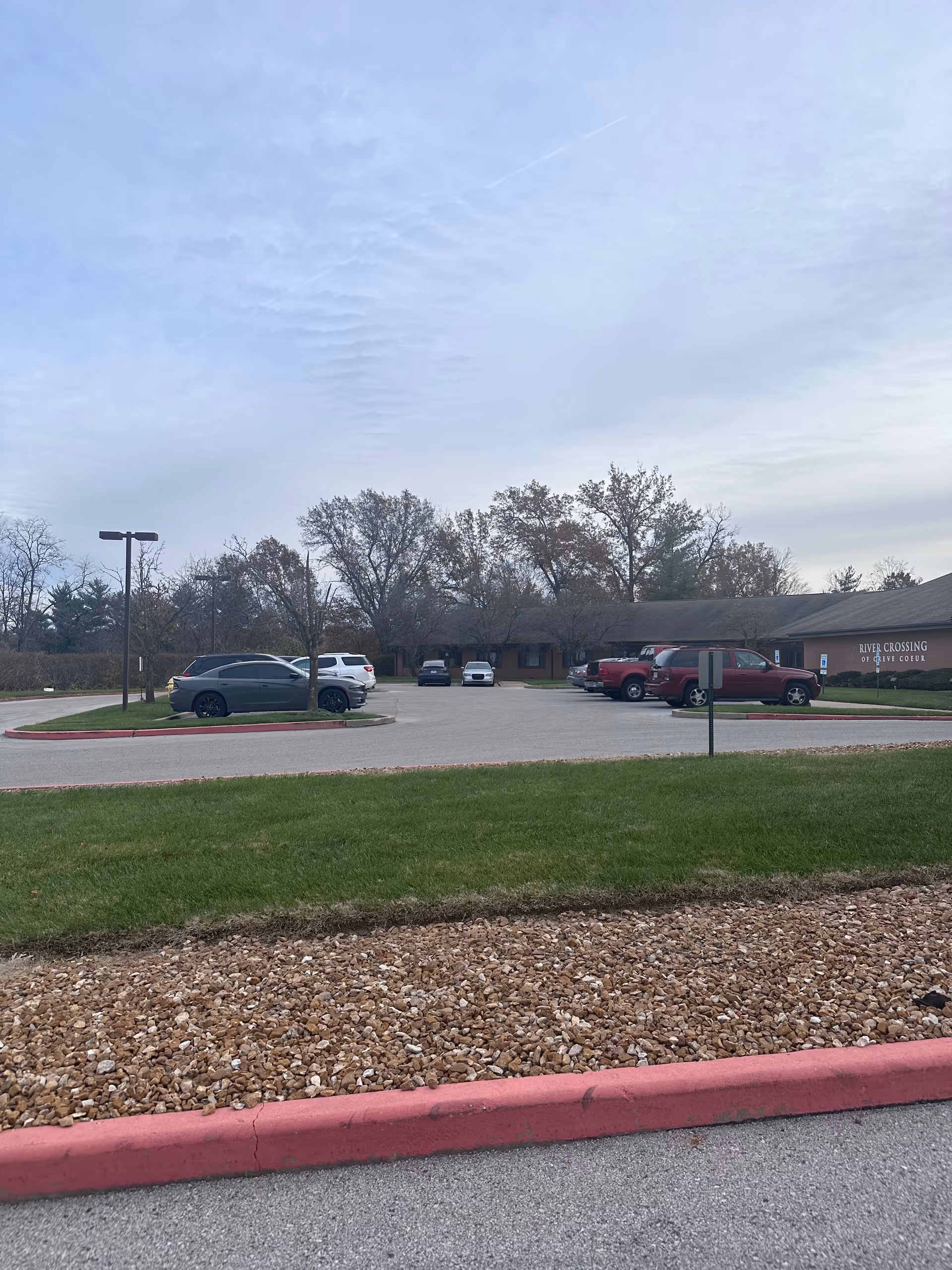 Front parking lot and entrance of River Crossing Rehab and Healthcare Center with parked cars, grass, and a cloudy sky.