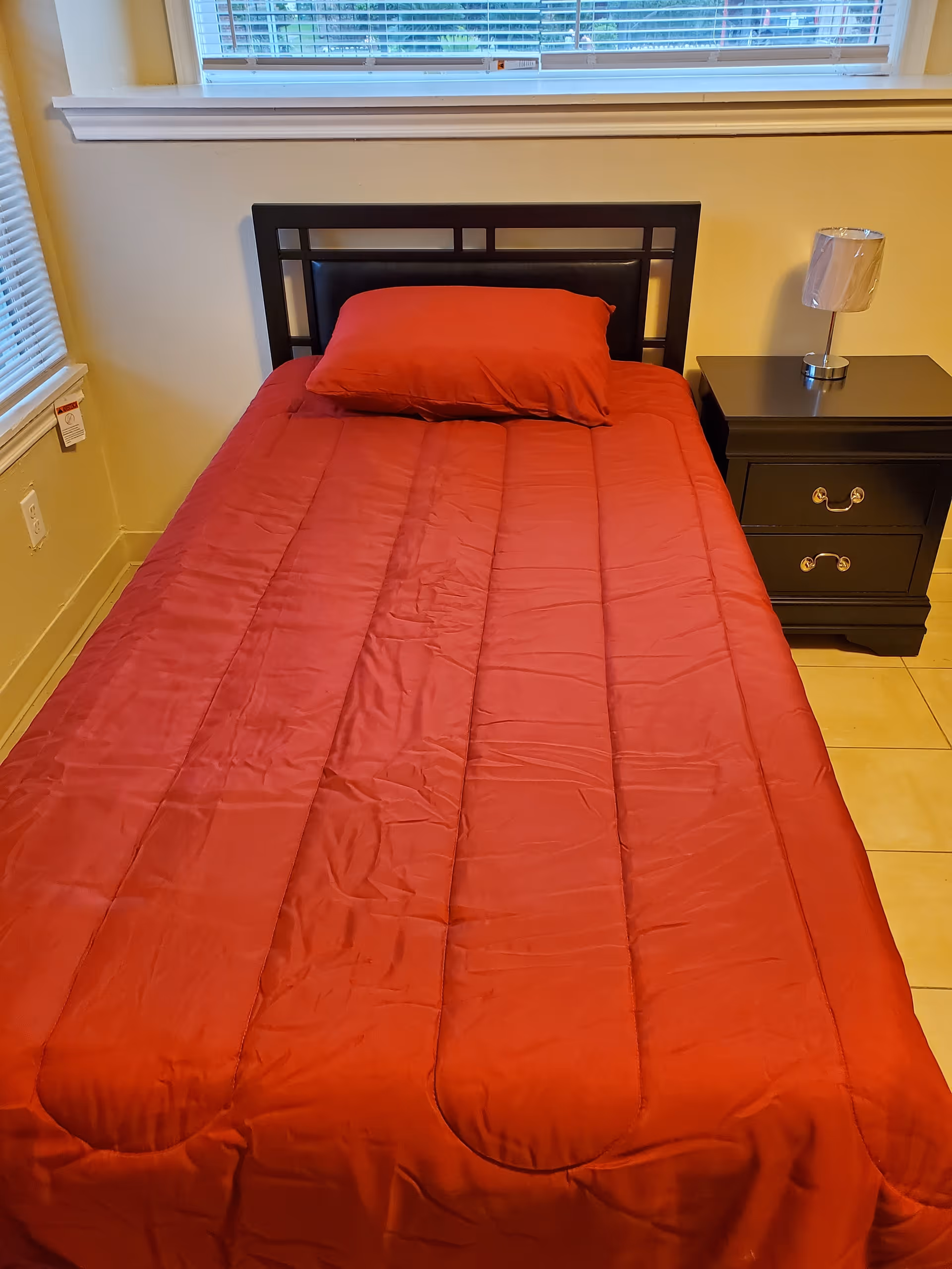 A single bed with a red comforter and matching pillow, a black headboard, and a black nightstand with two drawers and a small lamp on top. The room has beige walls and a window with blinds above the bed.