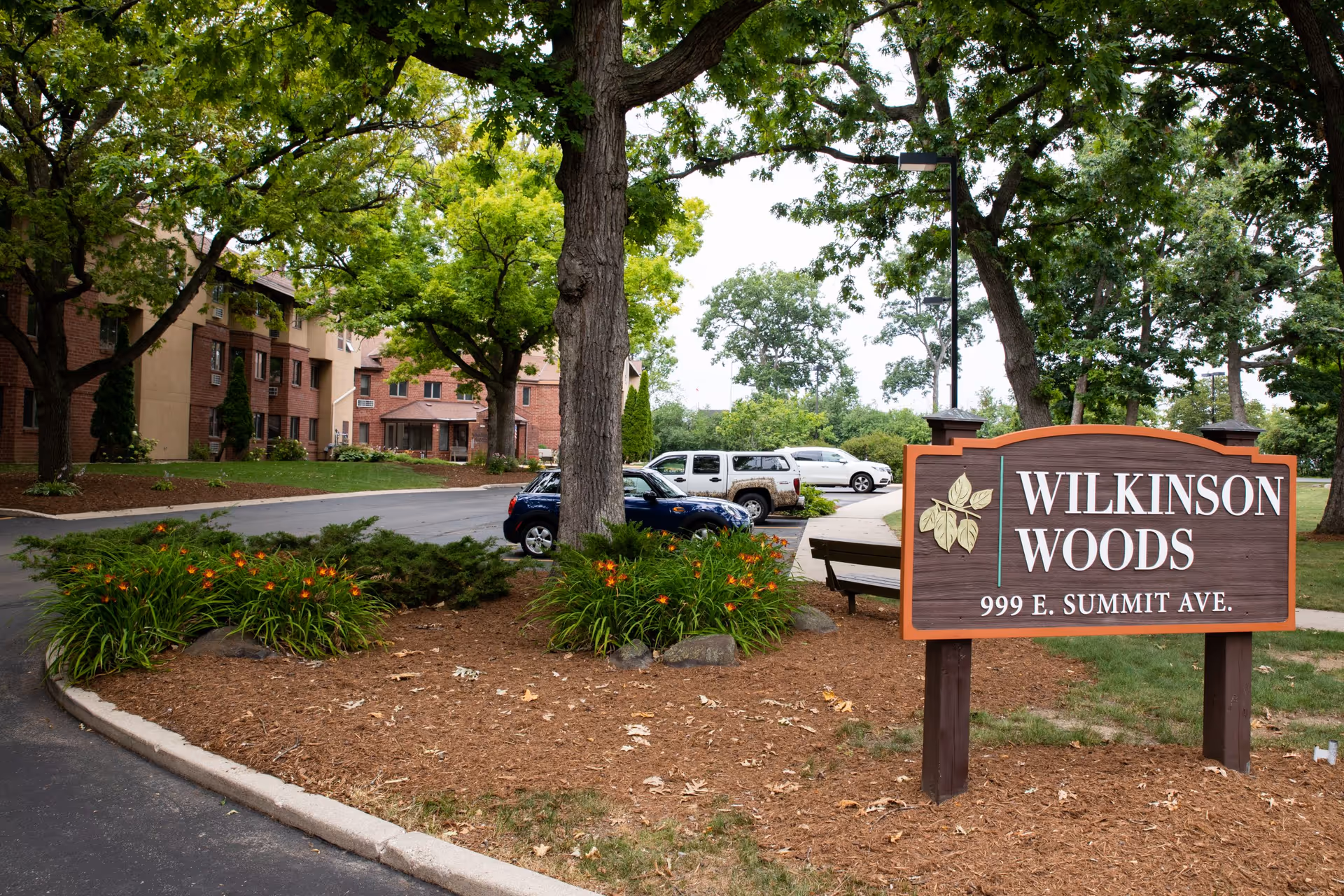Outdoor view of Wilkinson Woods Senior Community featuring a wooden sign with the community name and address, surrounded by trees, flowers, and parked cars near a multi-story brick building.