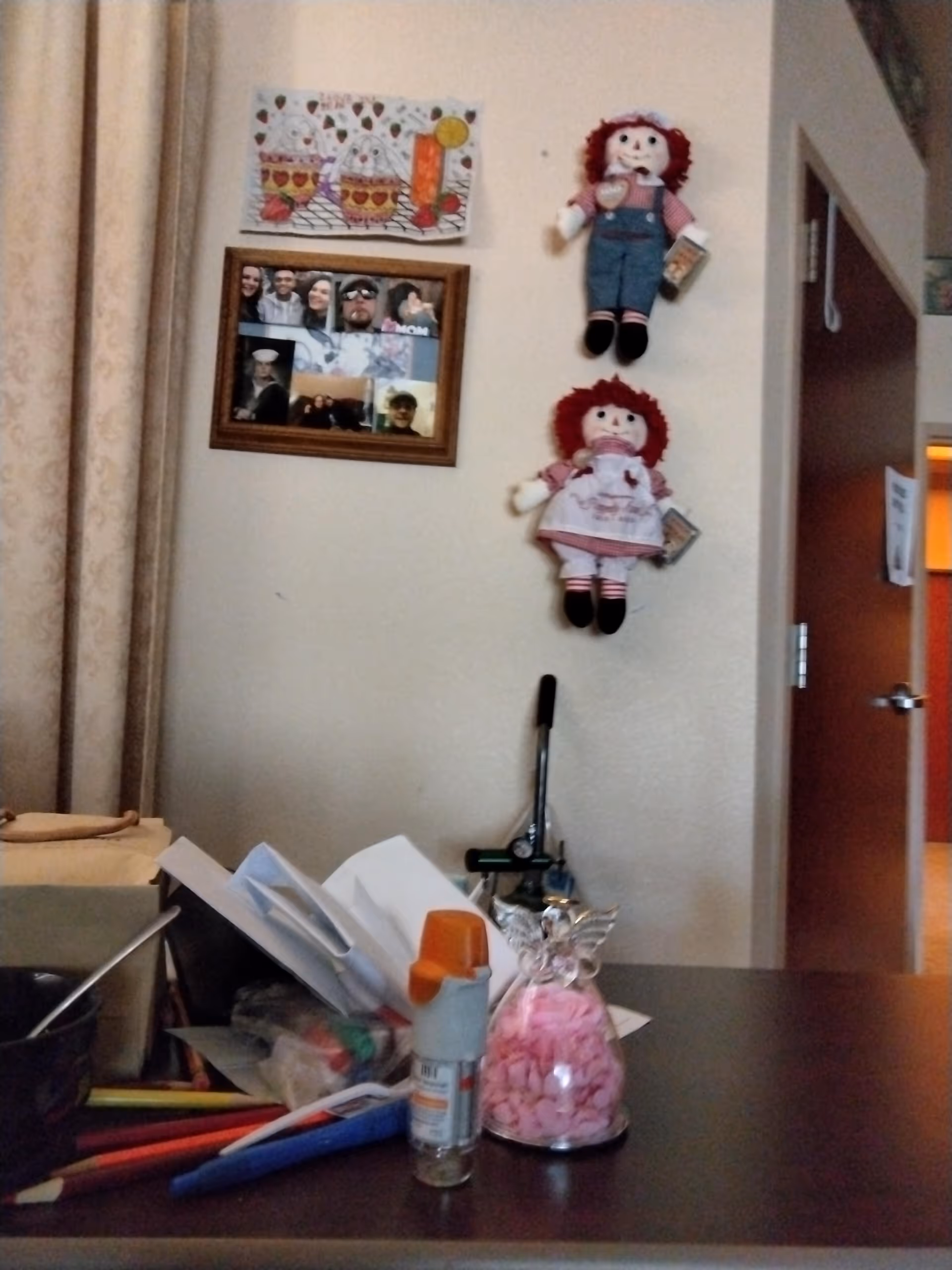 Interior office-like room with desk clutter, wall dolls, framed photos, and doorway to another area.