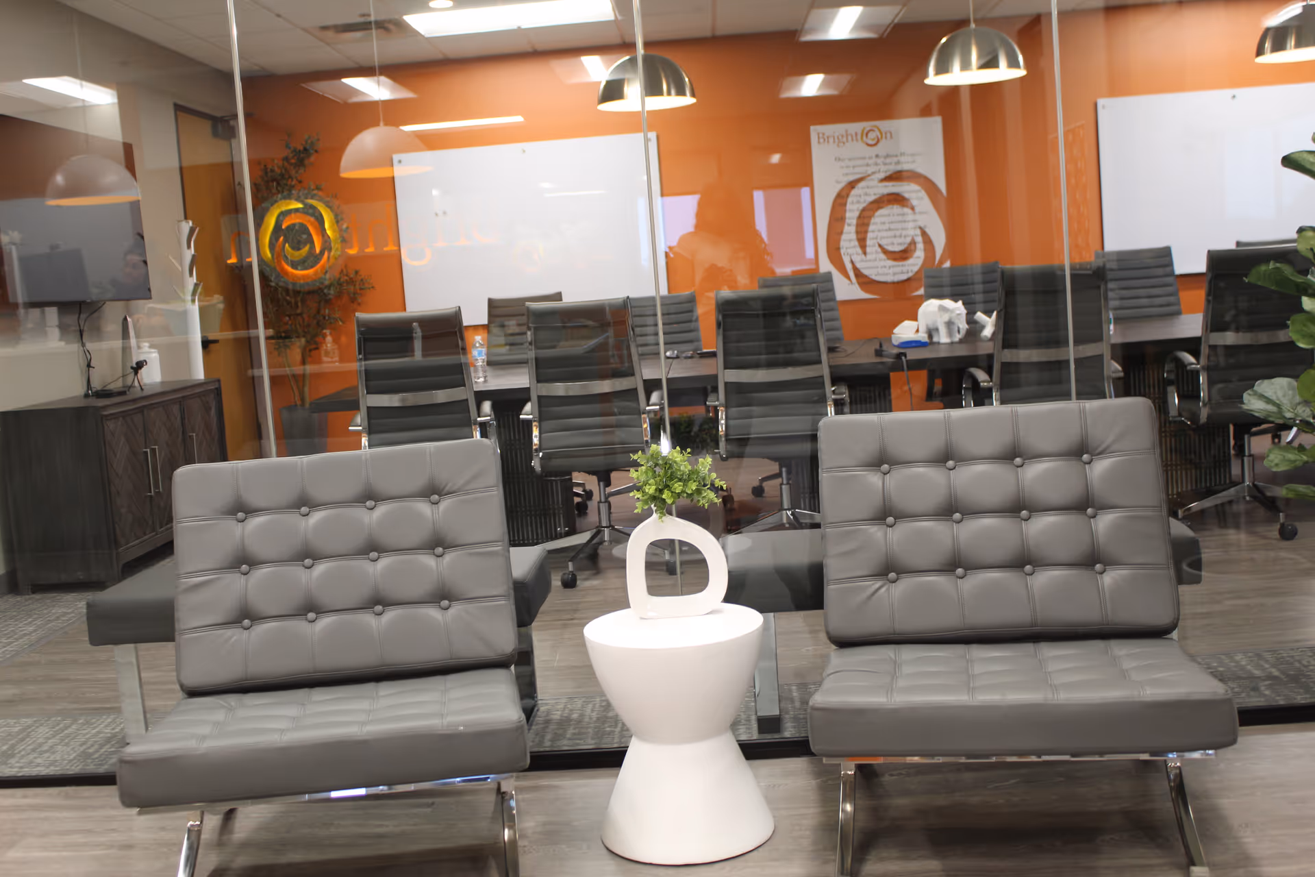Modern facility lobby with two gray tufted chairs, a white side table, plant, and glass walls.