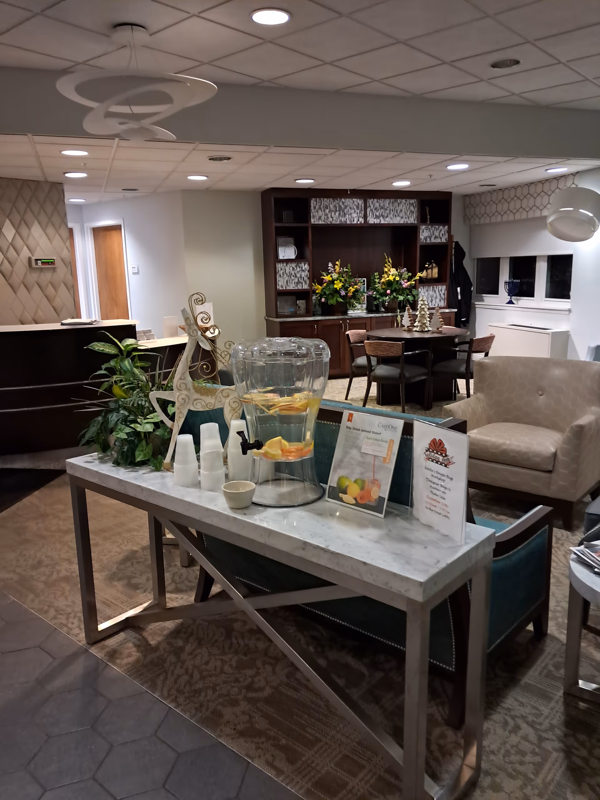 Indoor senior living lounge with a marble-topped table, water pitcher, plants, and seating.