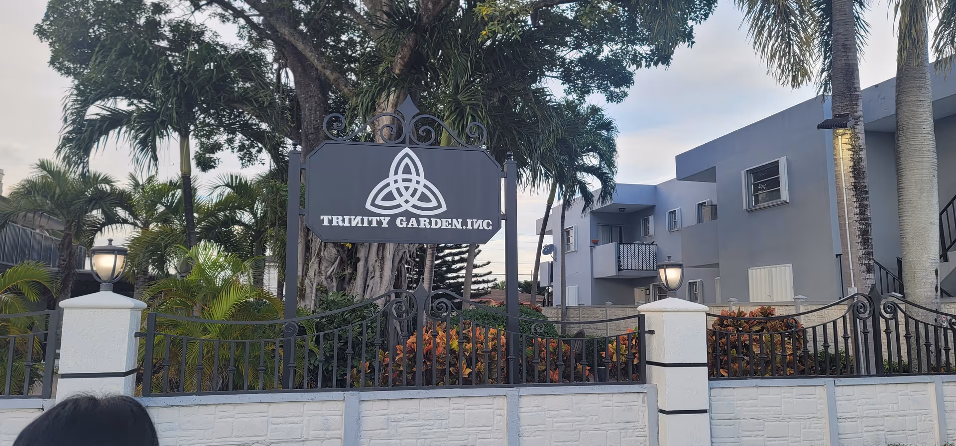 Exterior view of a gated entrance with a sign and tropical landscaping.