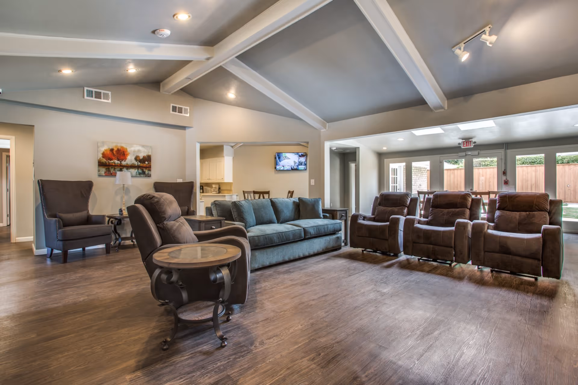 A spacious living room area with a vaulted ceiling and exposed beams. The room features a teal sofa, three brown recliners, two dark gray armchairs, and a round side table with a glass top. There is a painting of trees on the wall and a small lamp on a side table between the armchairs. In the background, a kitchen area and a dining table with chairs are visible. Large windows and glass doors let in natural light and provide a view of a fenced outdoor space.