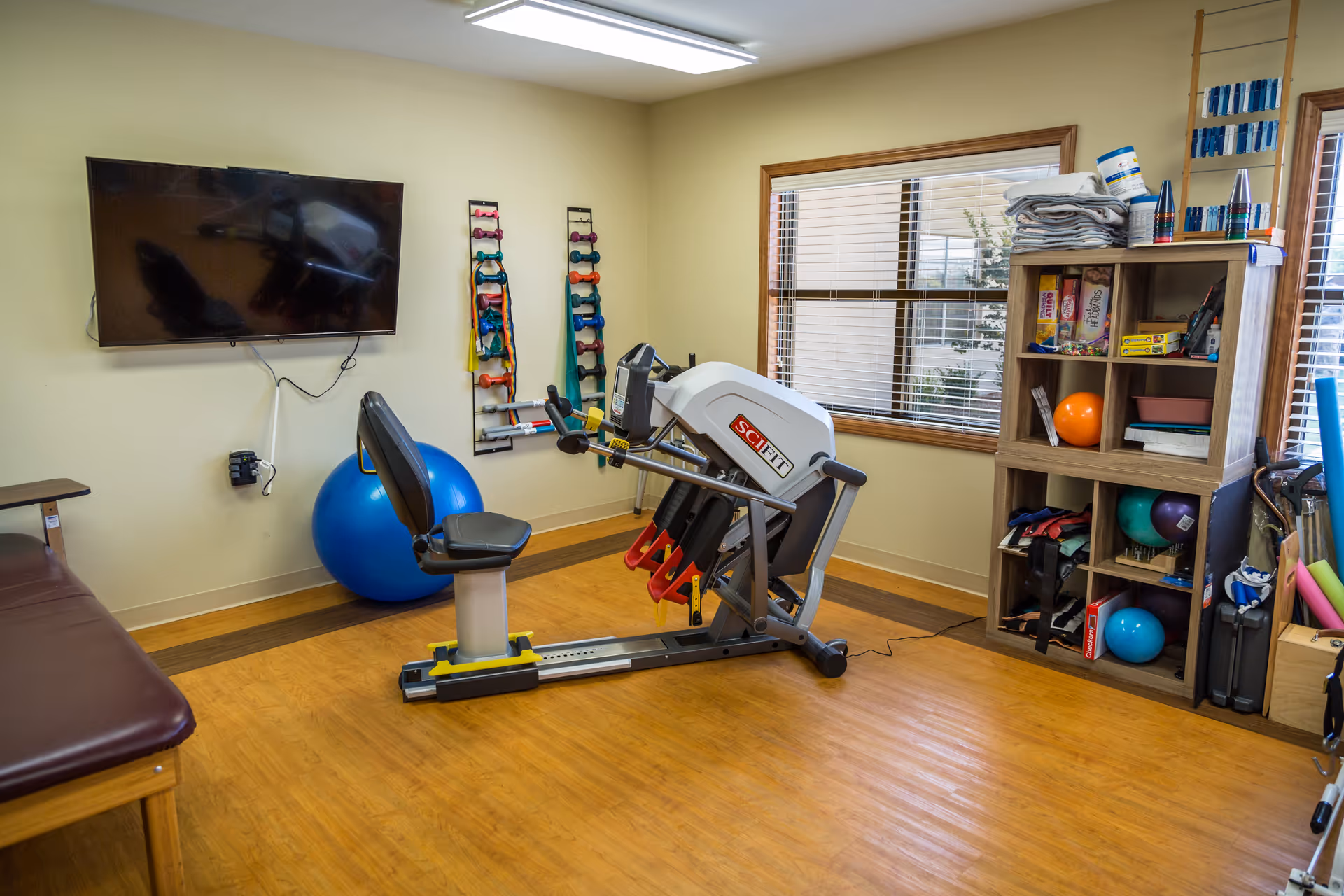 Rehab gym with treadmill, weights, shelves, and a mounted TV inside a care facility.