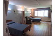Shared senior care room with two beds, curtains, chairs, and a large window.