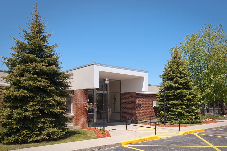 Exterior view of Medilodge of Rogers City with brick entrance, glass doors, and trees.