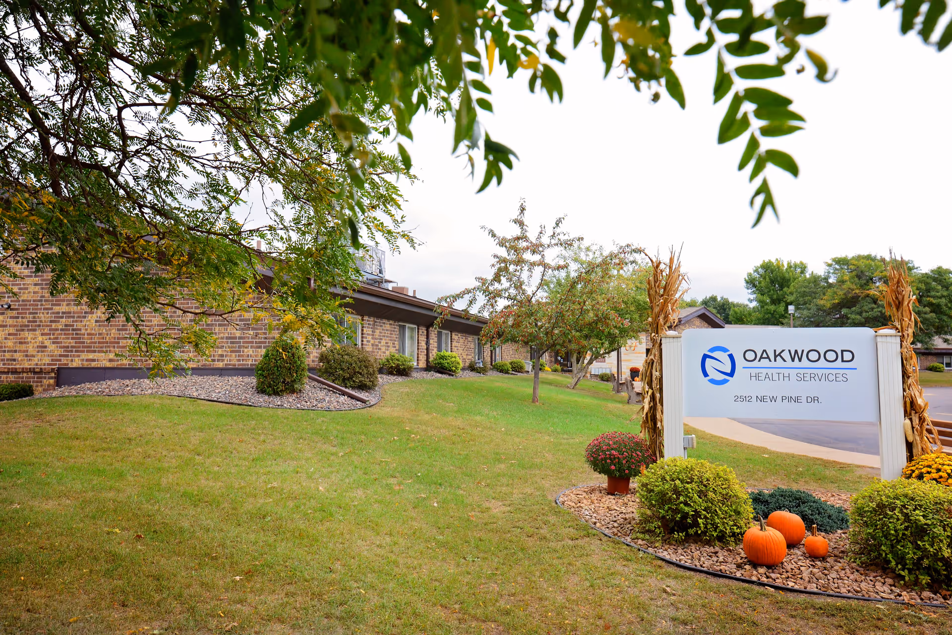 Single-story brick building and landscaped lawn with an Oakwood Health Services sign and seasonal pumpkins near the driveway.