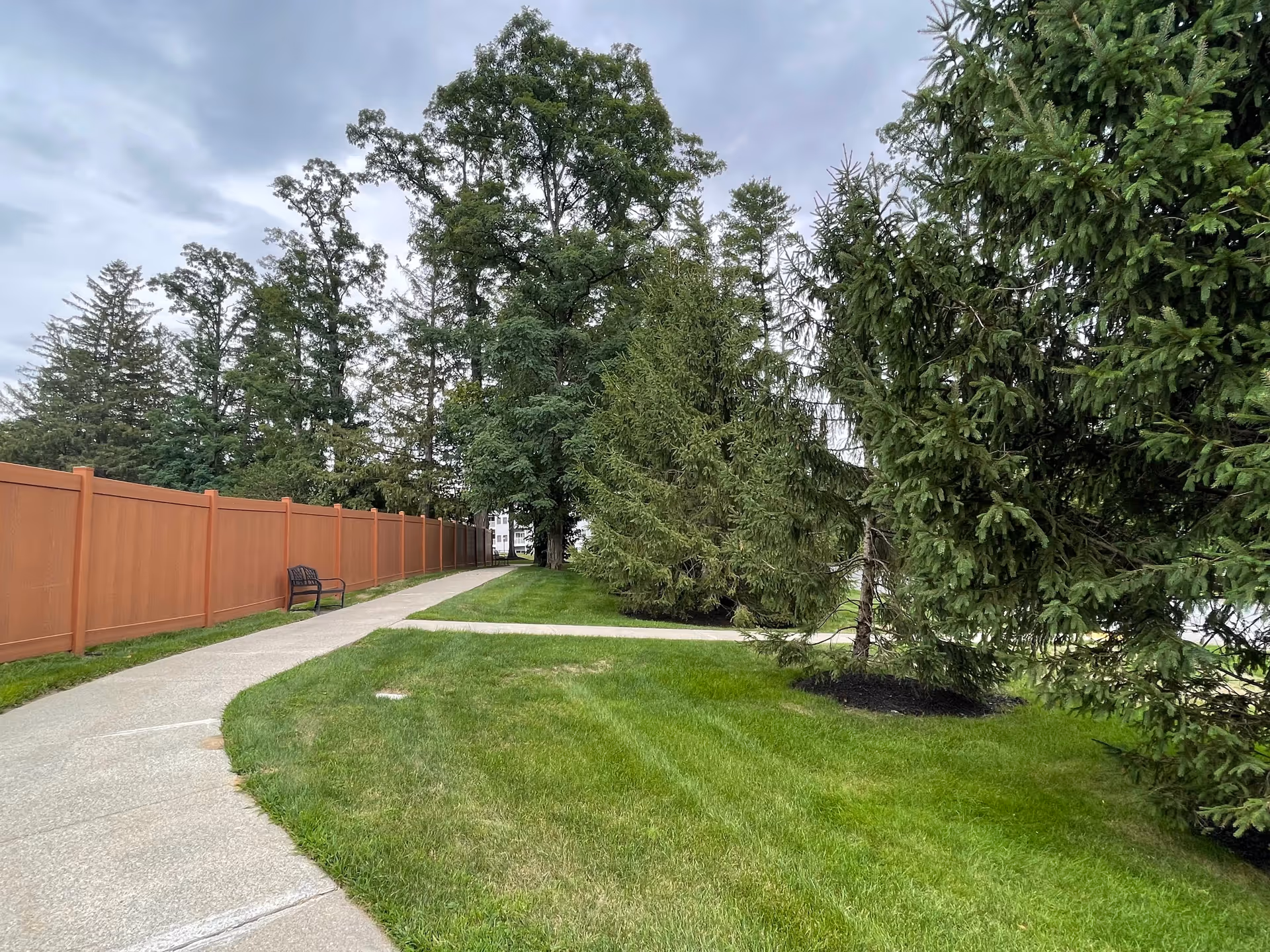 A paved walking path bordered by a brown fence on the left and green grass on both sides, with several large evergreen trees and other trees in the background under a cloudy sky. A bench is placed along the path near the fence.