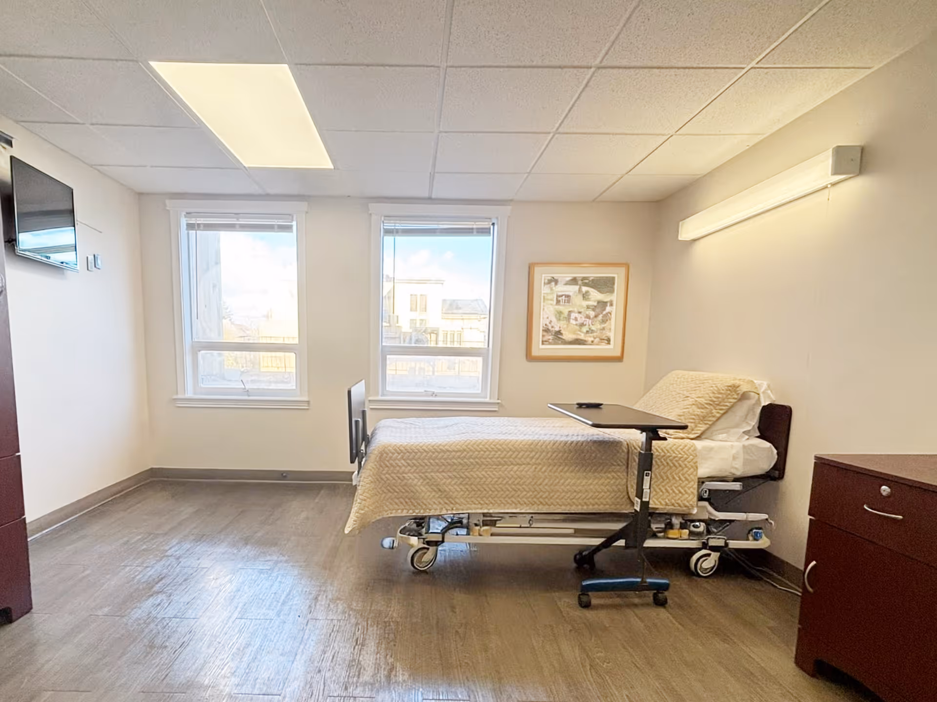 A bright and clean bedroom in a senior living facility with a hospital-style bed covered in a beige quilt, a small overbed table, two large windows letting in natural light, a wall-mounted TV, a framed picture on the wall, and wooden furniture including a dresser and a cabinet.