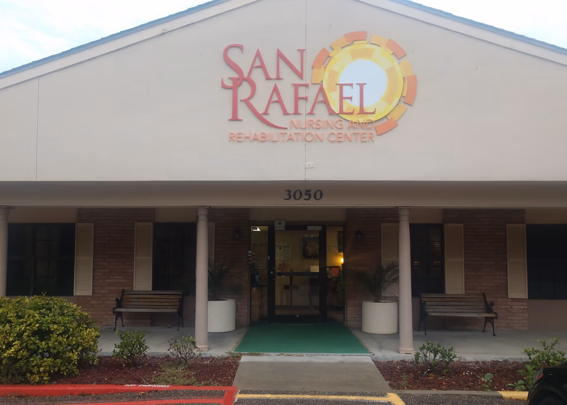 Front exterior view of San Rafael Nursing and Rehabilitation Center building entrance with two benches, potted plants, and the address number 3050 above the door.