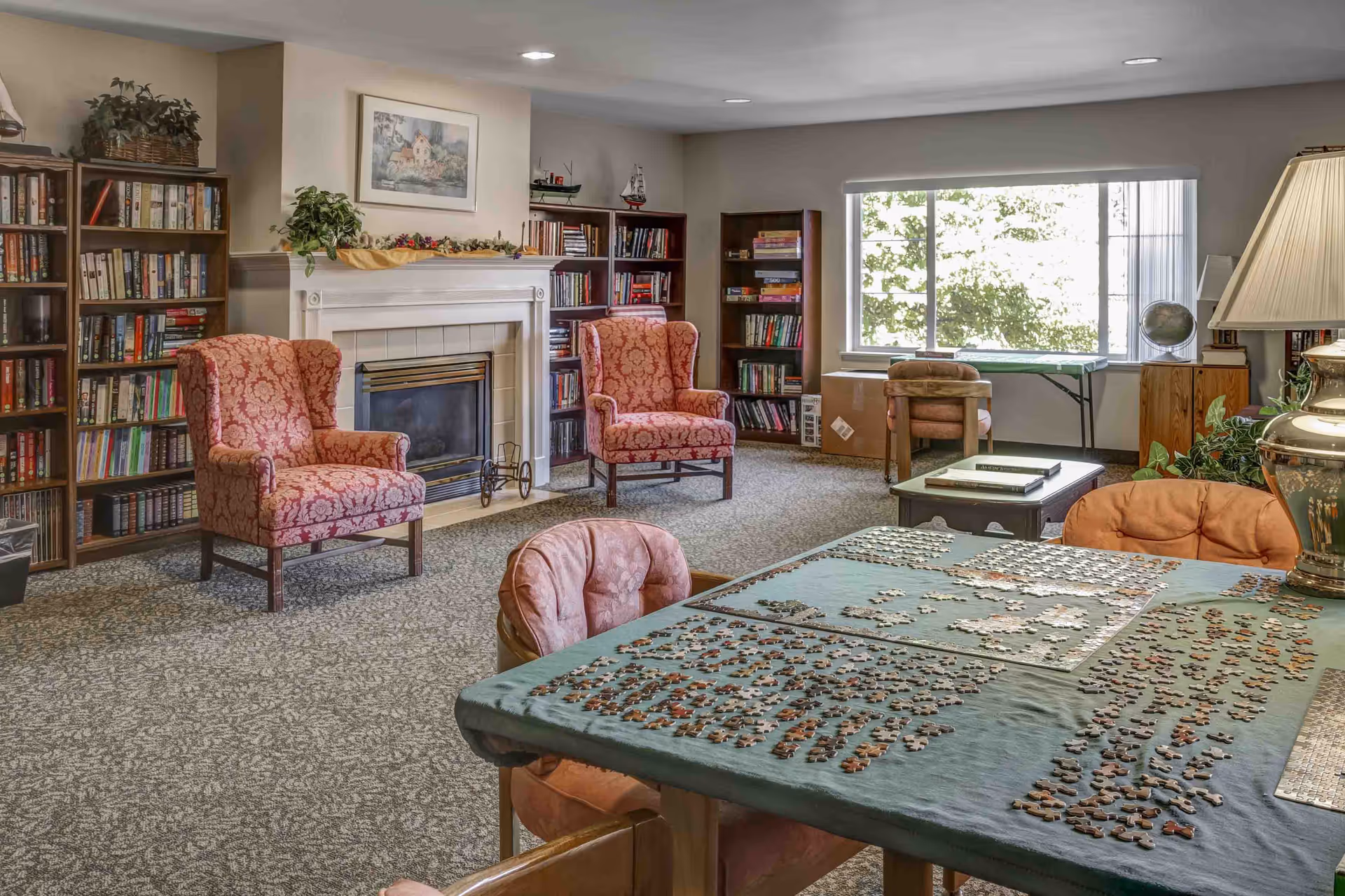 Bright common room with a fireplace, bookshelves, upholstered armchairs, and a table covered in jigsaw puzzle pieces.