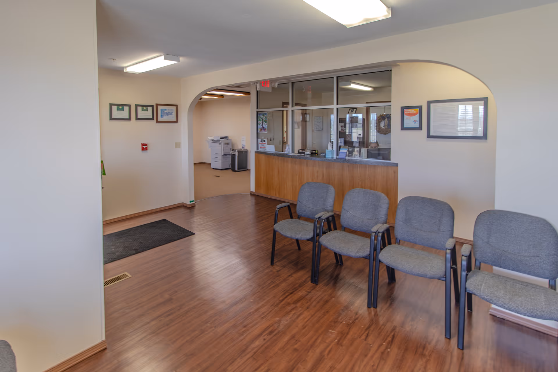 Indoor lobby with six gray chairs, a wooden floor, and a reception desk.