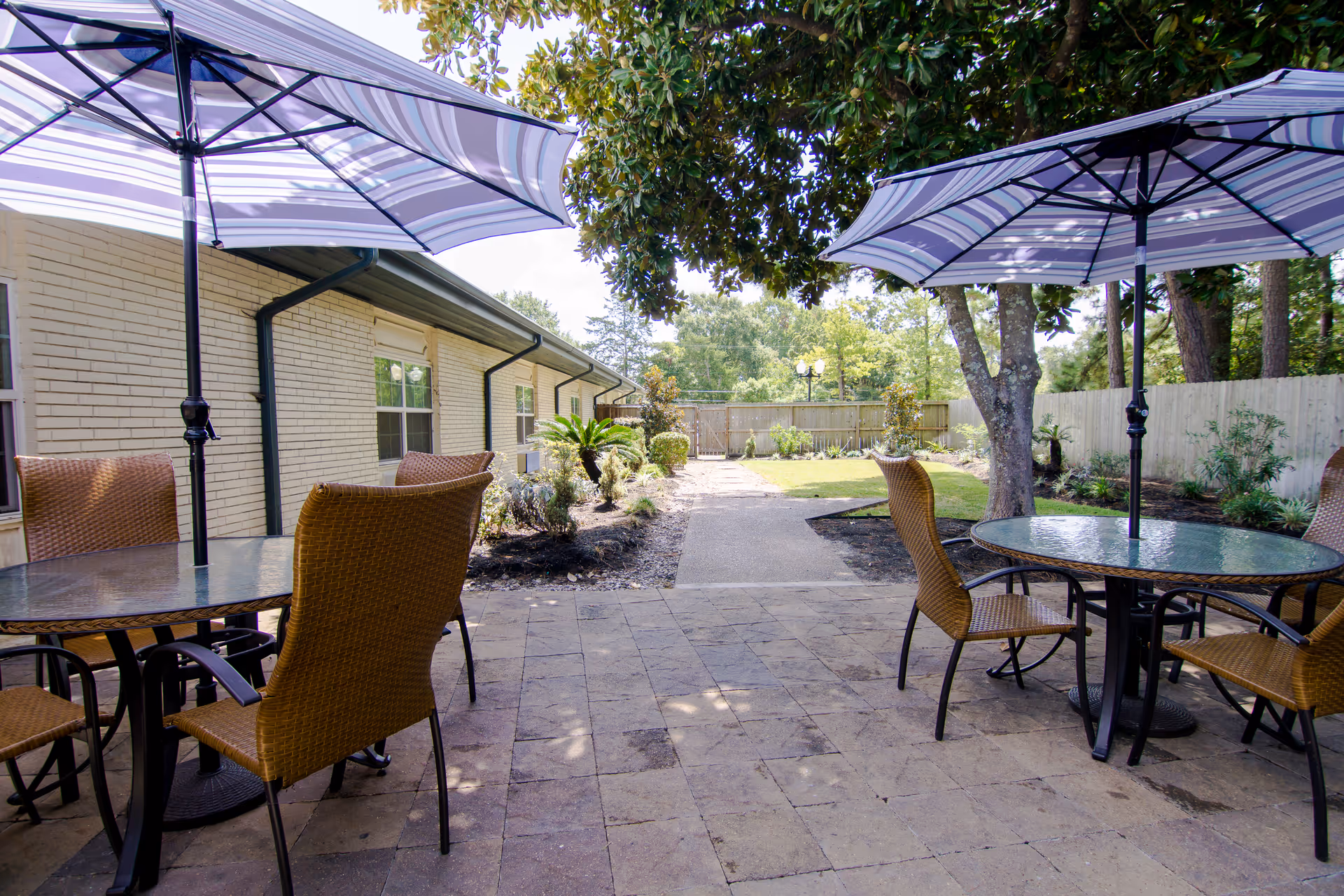 Outdoor patio area with two round glass tables, each shaded by a large striped umbrella. Four wicker chairs are arranged around the tables. The patio is paved with stone tiles and is adjacent to a light-colored brick building. There are trees, plants, and a fenced garden area in the background.