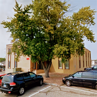 Exterior view of a beige brick building with a large tree and parked cars.