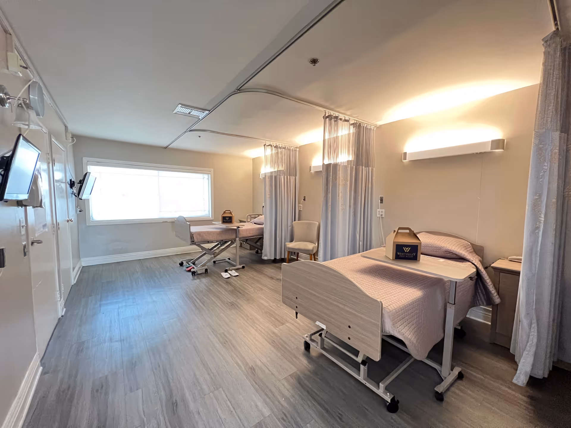 Two hospital-style beds with curtains in a bright shared patient bedroom.