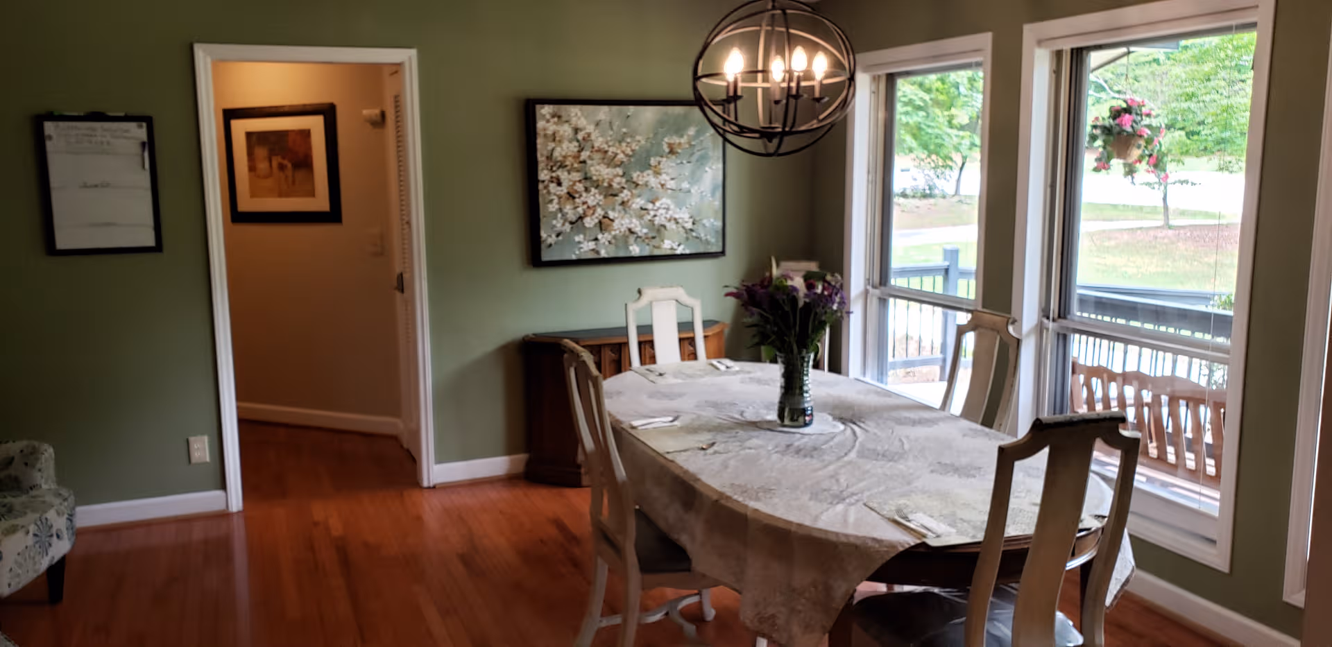 Dining room with an oval table, chairs, flowers, green walls, and large windows.