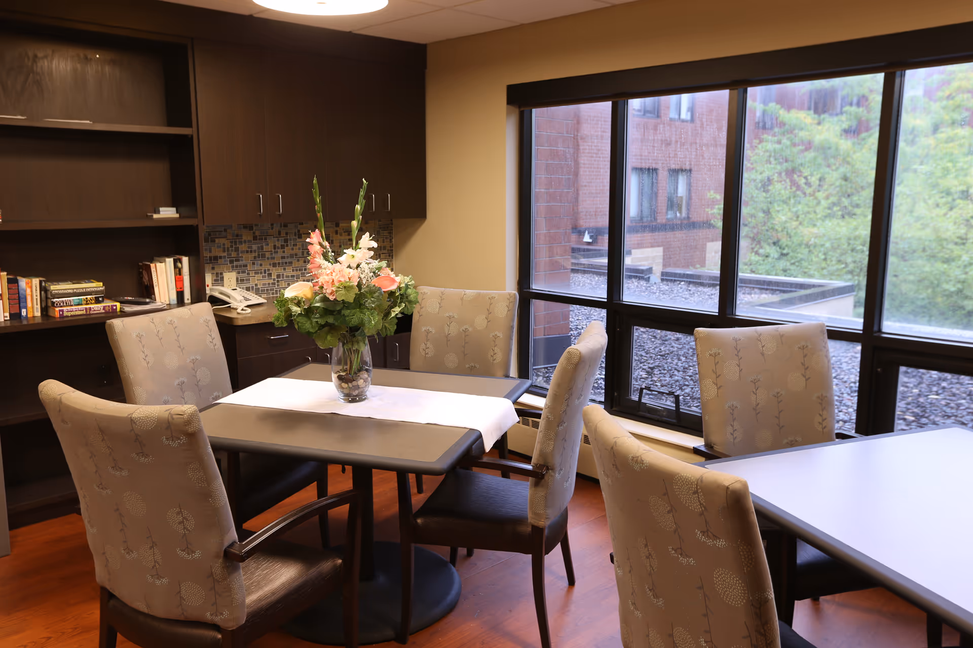 Interior dining area with tables, upholstered chairs, and a flower centerpiece near large windows.