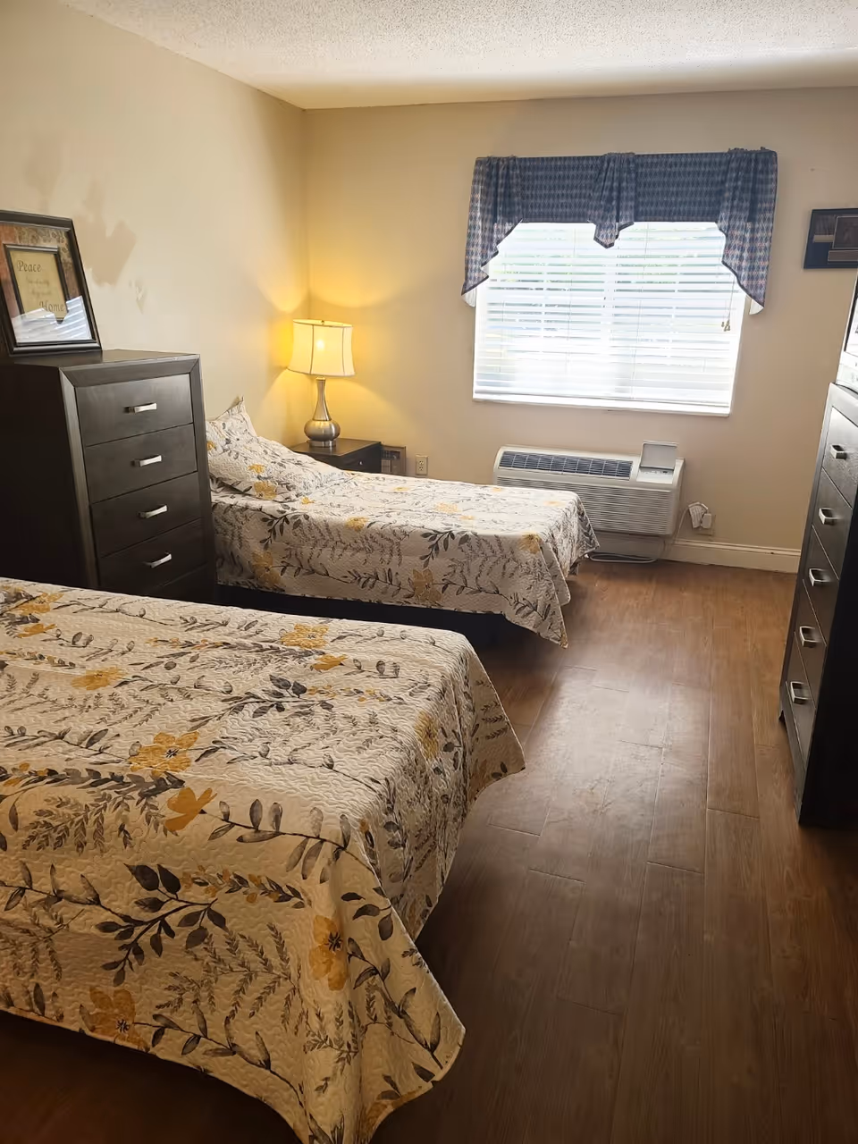 Two-bed furnished bedroom with matching floral bedspreads, dressers, a bedside lamp, and a window with an air conditioner.