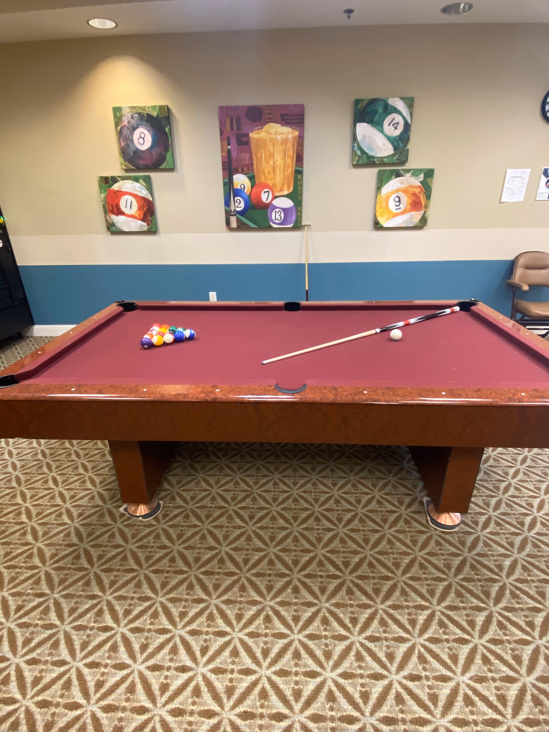 Indoor room with a burgundy pool table set up for a game, including a cue stick and a rack of billiard balls. The wall behind the table has five colorful paintings featuring billiard balls and a glass of iced drink. The floor is carpeted with a geometric pattern, and there is a chair and some papers on the wall to the right.