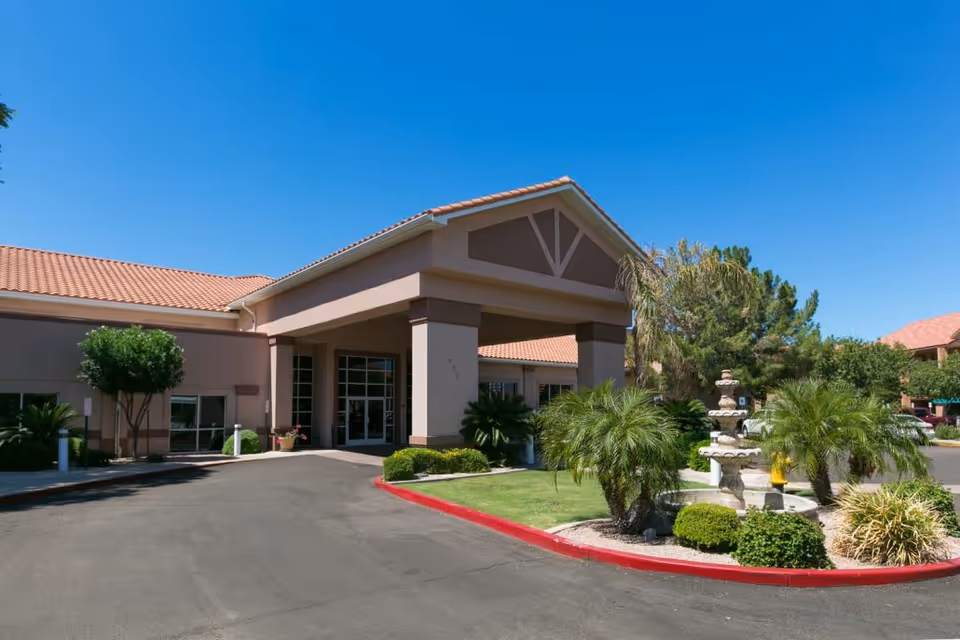 Senior living facility entrance with covered porte-cochere and landscaped driveway under blue sky.