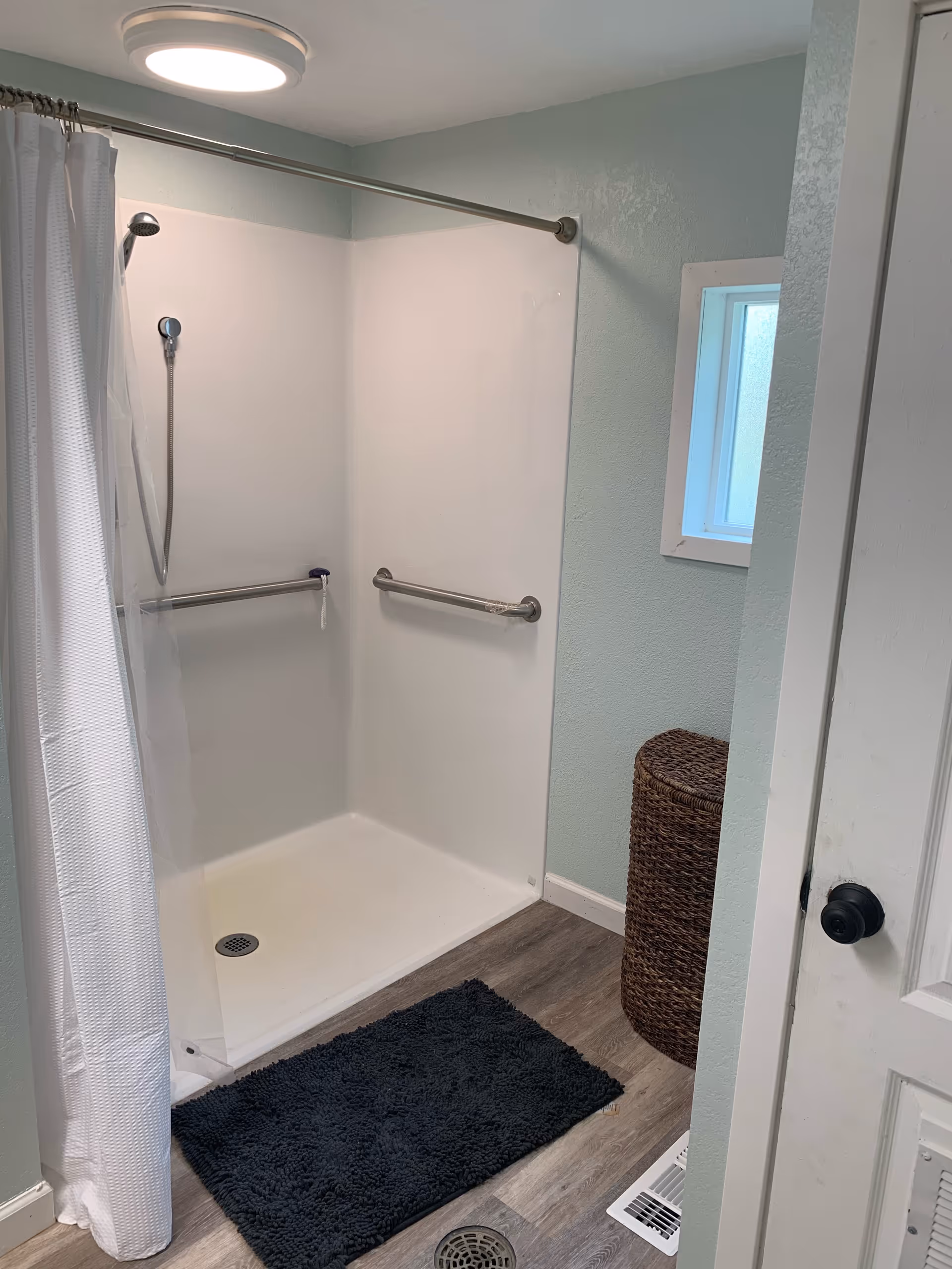 Shower stall with grab bars, curtain, and a small window in a light blue bathroom.