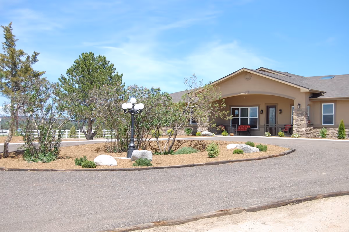 Exterior view of a single-story assisted living facility building with a covered porch, outdoor seating, and a circular driveway surrounded by landscaped bushes, rocks, and a vintage-style street lamp under a clear blue sky.