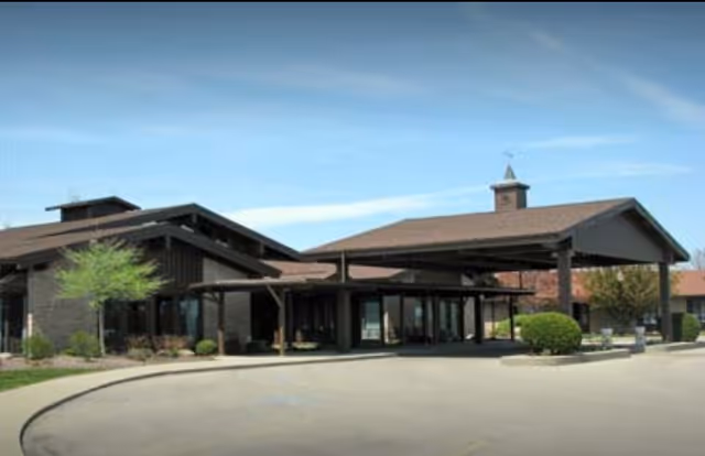 Front entrance of a one-story retirement community building with a covered porte-cochère, glass doors and landscaped shrubs.