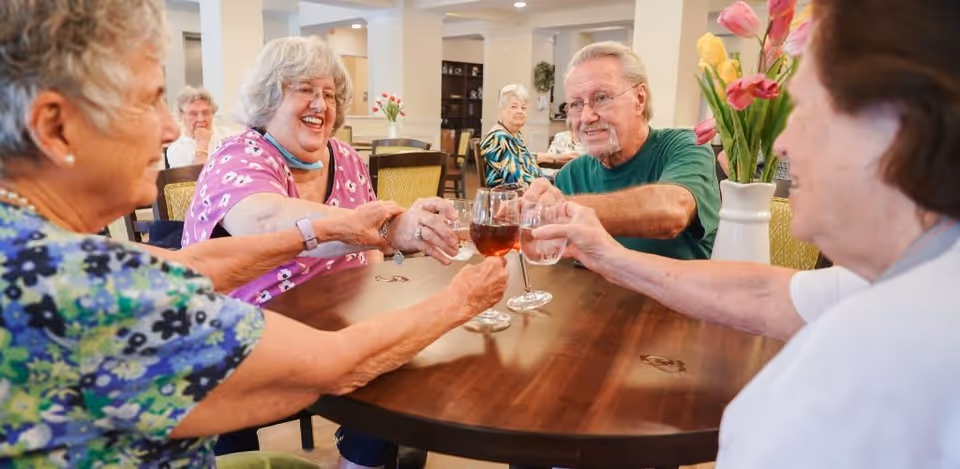 Four elderly people sitting around a wooden table in a well-lit room, smiling and clinking their glasses together in a toast. The room has light-colored walls, decorative flowers on the table, and other people seated in the background.