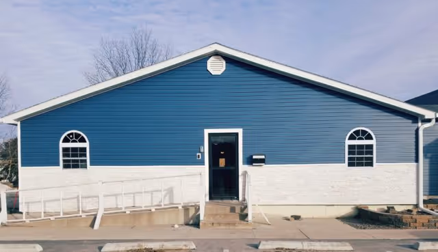 Exterior front view of a single-story building with blue siding and white lower walls. The building has a central door with a small set of stairs and a wheelchair ramp leading up to it. There are two arched windows on either side of the door. The sky is clear with some light clouds.