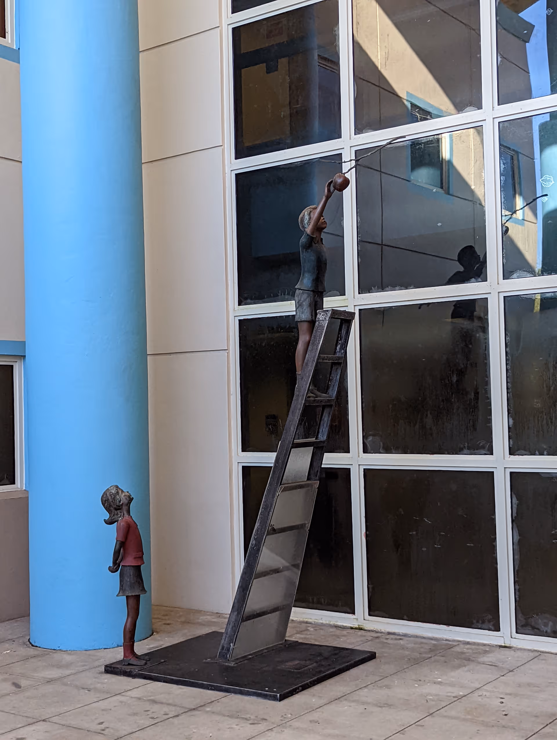 Exterior of a building with large glass windows, blue column, and child sculptures beside a ladder
