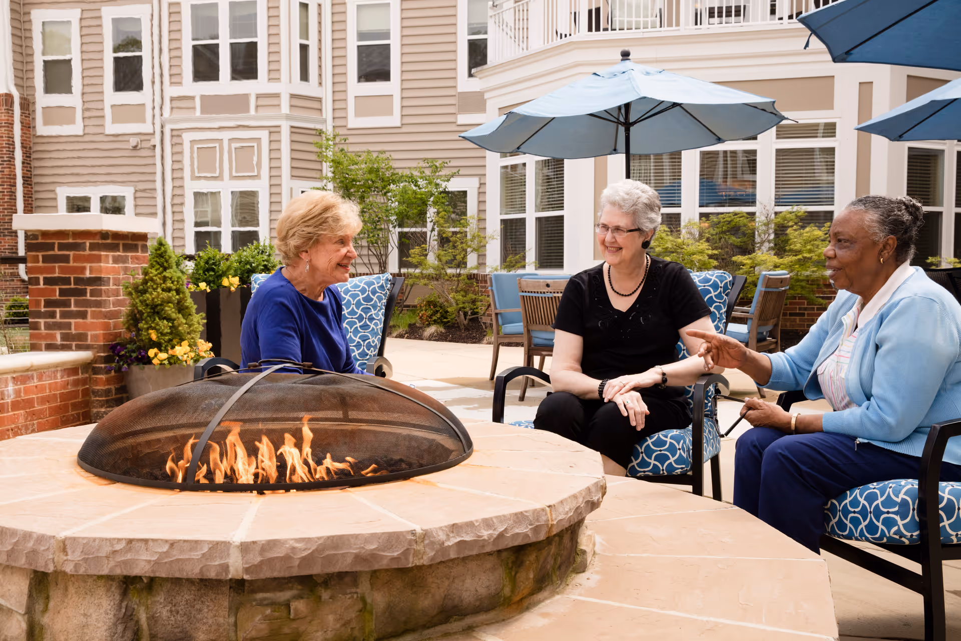 Three elderly residents chat around a lit fire pit on Brightview Severna Park's outdoor patio.