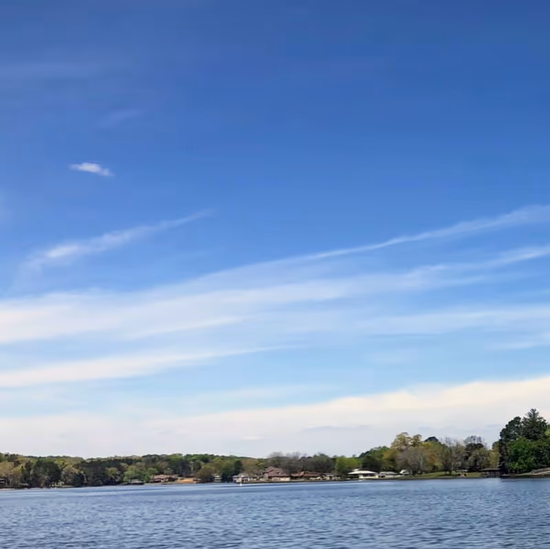 Calm lake with trees and houses along the shoreline under a blue sky.
