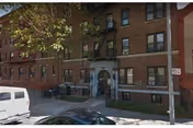 Brick apartment-style building facade with arched doorway and parked cars along a tree-lined street.