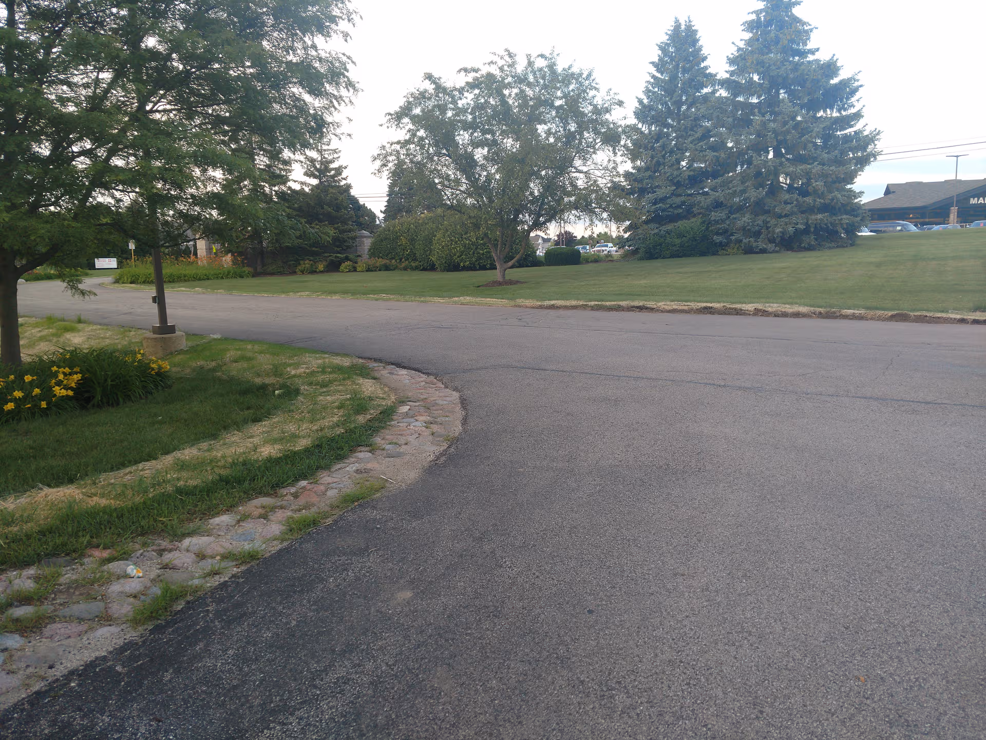 Curved road bordered by green lawns and trees leads toward a distant building.
