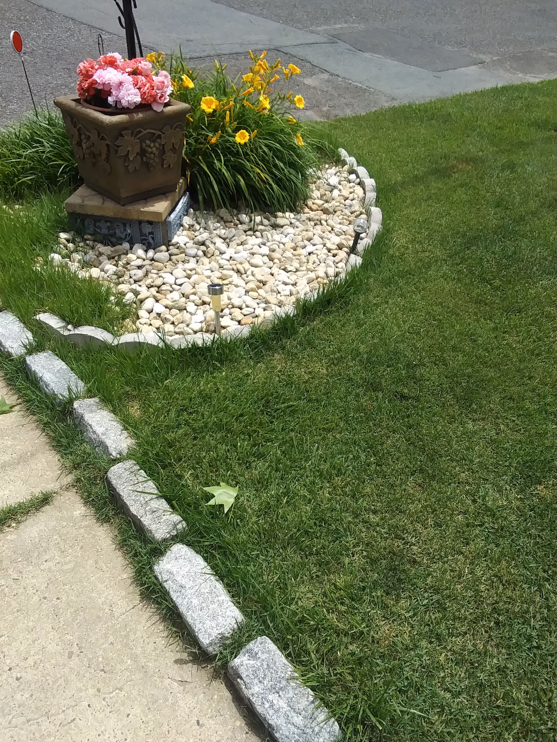 Outdoor garden bed with a decorative planter on a pedestal, surrounded by rocks and green lawn.