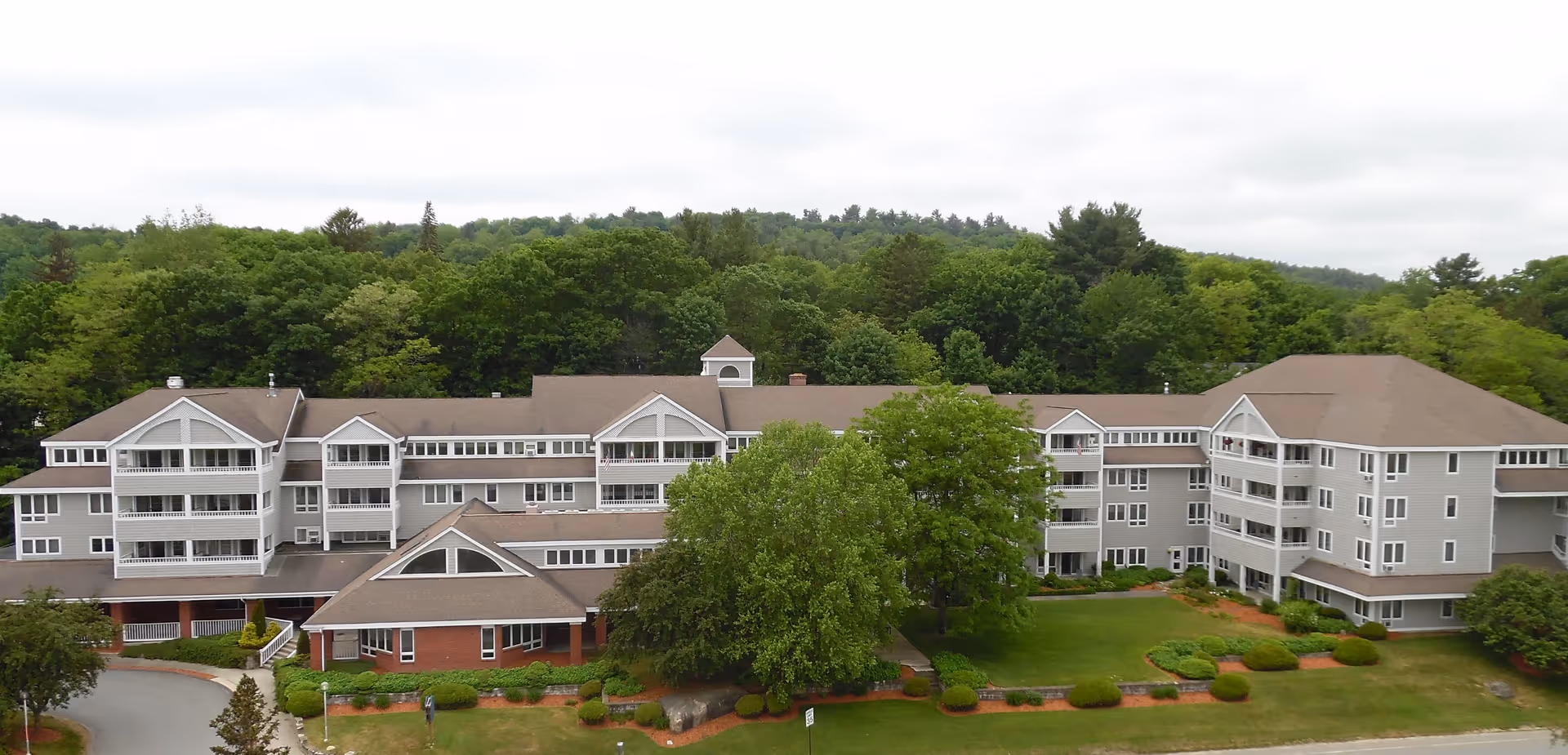 Exterior view of a large multi-story senior living facility building surrounded by green trees and landscaped lawns under a cloudy sky.